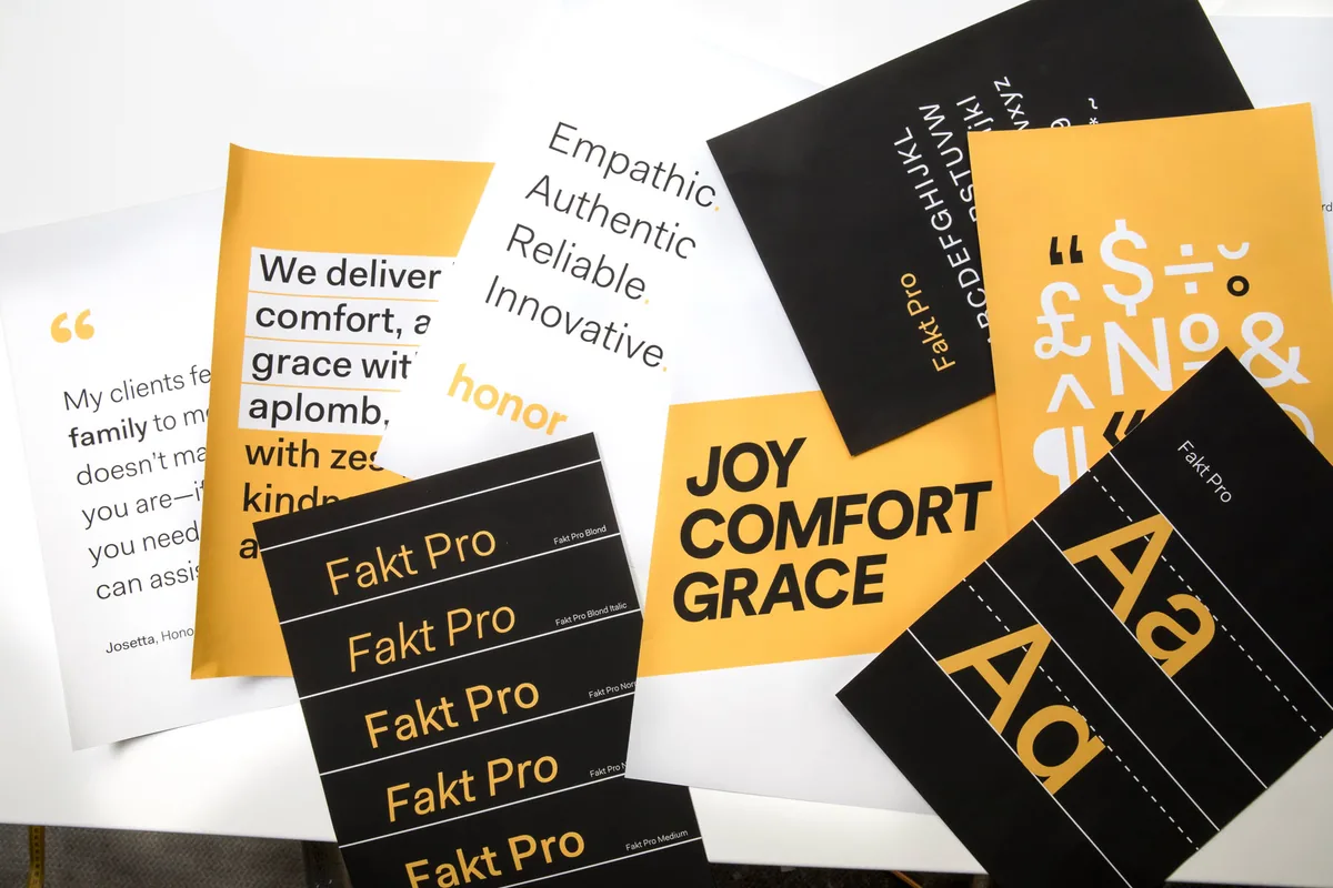 Printed Honor brand posters on a table: the honor wordmark, Fakt Pro type specimens, a quote card and the words joy, comfort, grace in black and amber